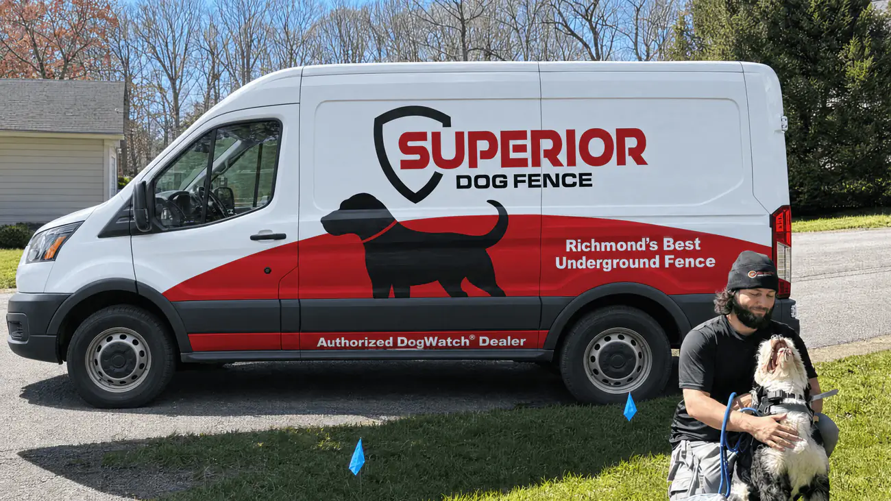 A dog owner kneeling with a malamute on a lawn marked with blue boundary training flags, beside the Superior Dog Fence van.