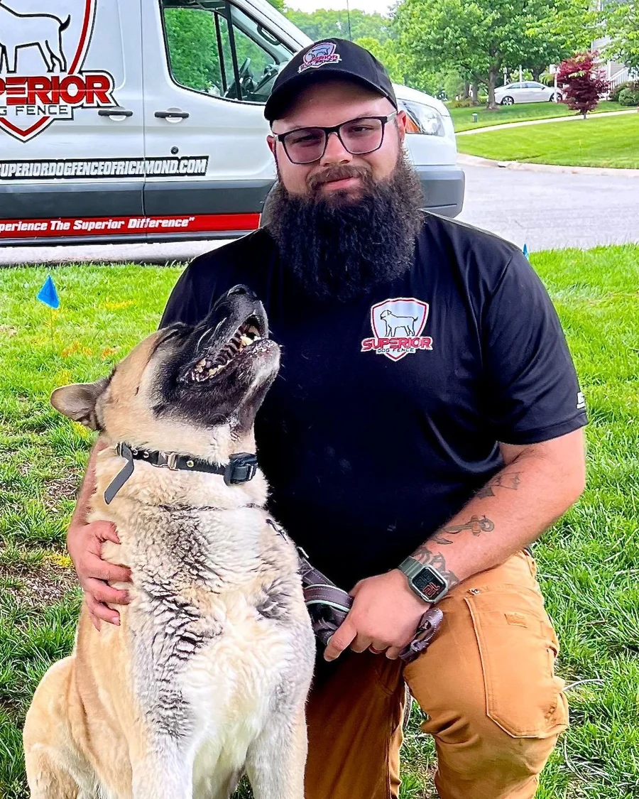 A Superior Dog Fence installer kneeling beside a dog in a front yard, with the company van behind and a boundary flag in the grass.