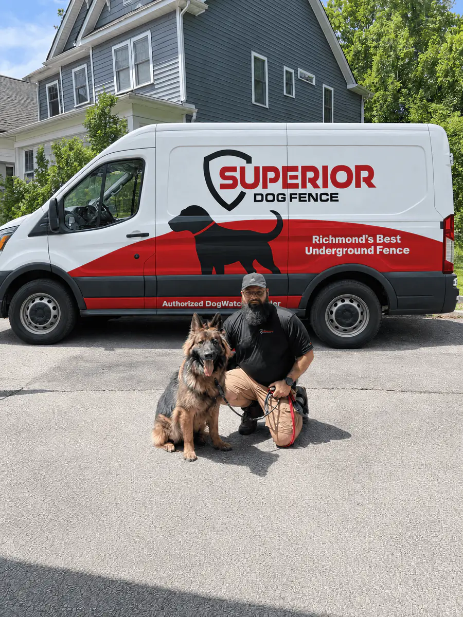 A Superior Dog Fence installer kneeling with a German shepherd sitting on the driveway beside the company van.