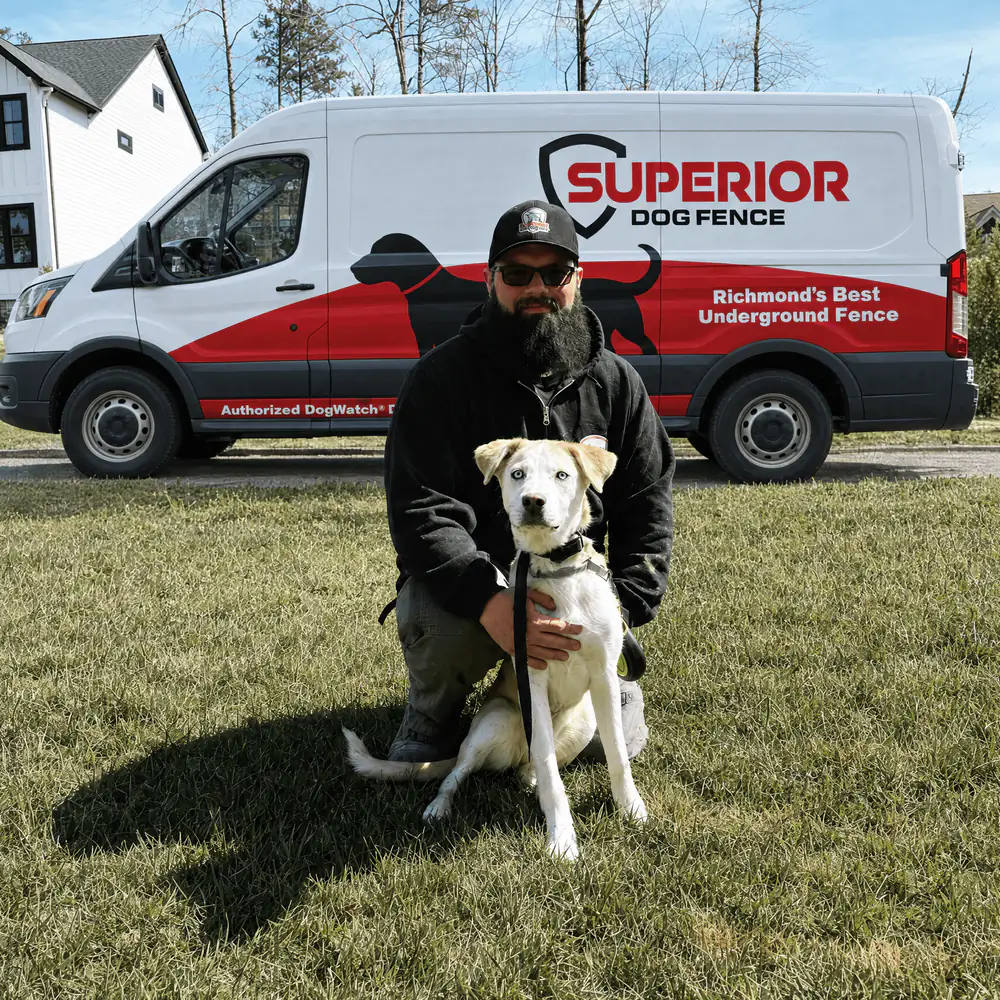 A Superior Dog Fence installer kneeling on the lawn with a white dog, the company van behind him.